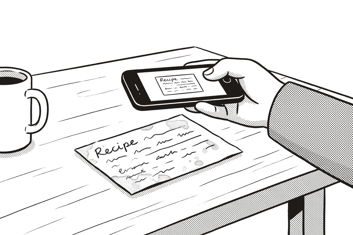 A hand holding a phone over a stained recipe card on the table, the card on the phone's screen.