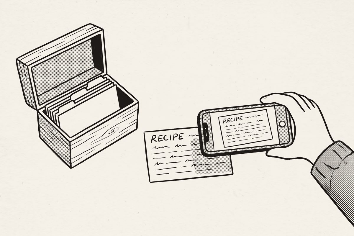 A hand holding a phone over a recipe card to photograph it, beside the open recipe box.