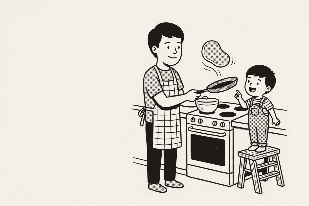 A dad flipping pancakes while his small son watches from a step stool.