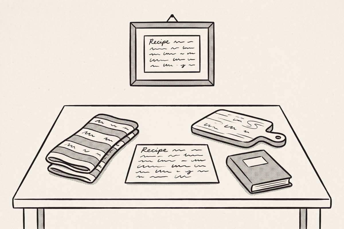 A recipe card on a kitchen table among a tea towel, a cutting board and a small cookbook, a framed recipe card on the wall above.