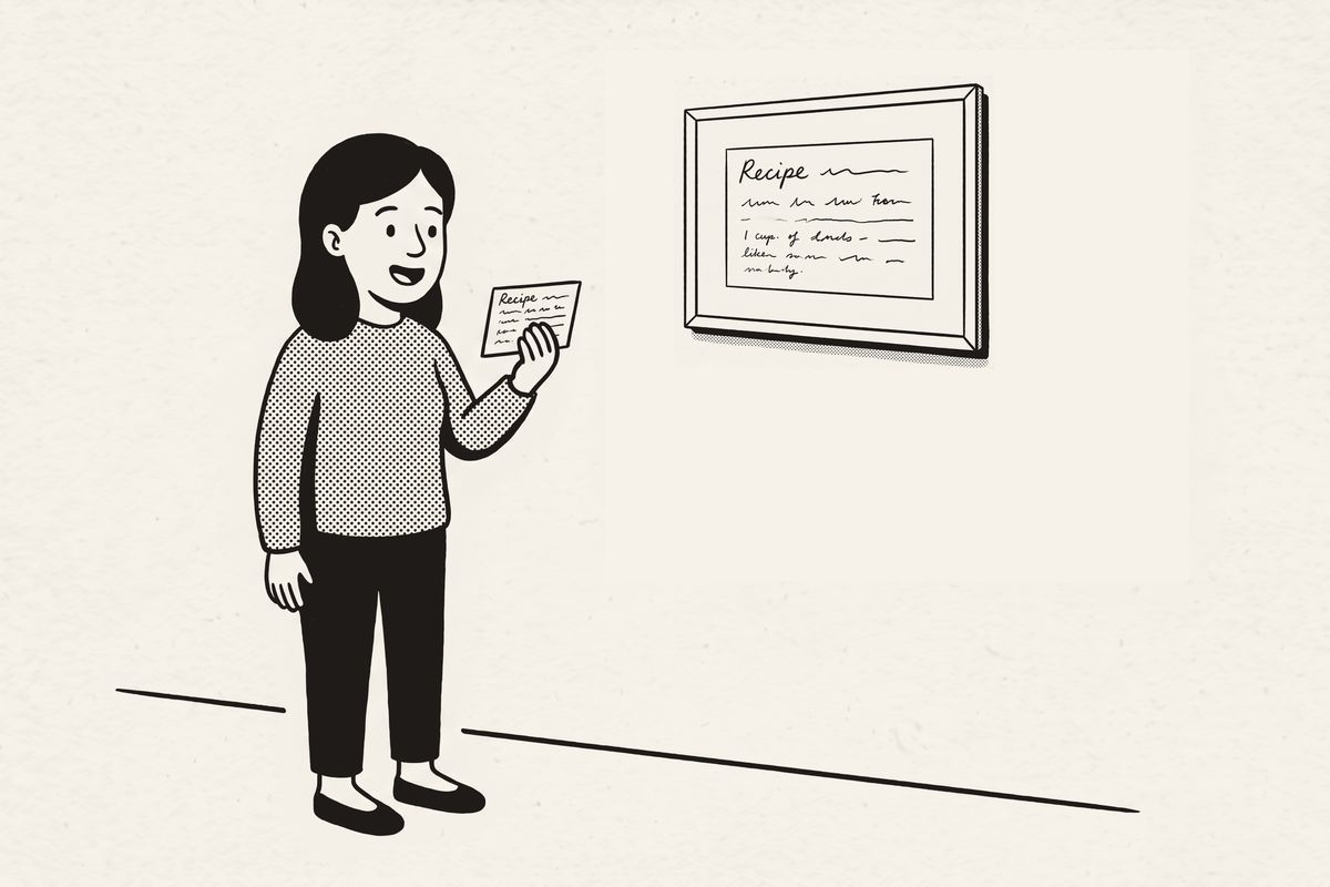 A woman holding up a small recipe card beside its large framed print on the wall.